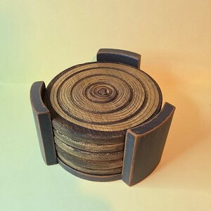Pu&ograve; includere: Una pila di sottobicchieri circolari in legno caratterizzati da un motivo a venature naturali ad anelli di crescita in tonalit&agrave; marrone e marrone chiaro. I sottobicchieri sono tenuti insieme da un supporto in legno grigio antracite scuro.