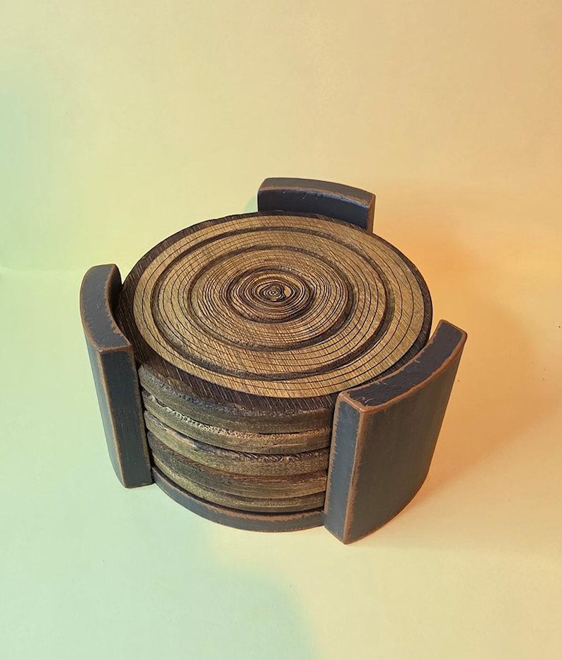 Pu&ograve; includere: Una pila di sottobicchieri circolari in legno caratterizzati da un motivo a venature naturali ad anelli di crescita in tonalit&agrave; marrone e marrone chiaro. I sottobicchieri sono tenuti insieme da un supporto in legno grigio antracite scuro.