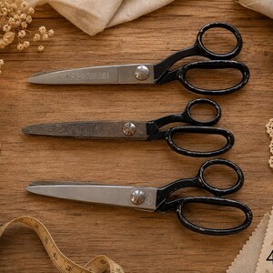May include: Three pairs of vintage scissors with black handles and silver blades are arranged on a wooden surface. The scissors are accompanied by sewing notions, including buttons, spools of thread, and a measuring tape. The image has a warm, rustic aesthetic.