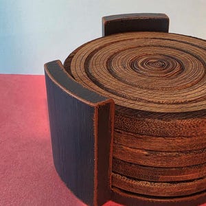 Pu&ograve; includere: Una pila di sottobicchieri circolari in legno con venature visibili, inseriti in un supporto in legno nero con finitura opaca. I sottobicchieri presentano anelli concentrici che ricordano i cerchi di crescita degli alberi.