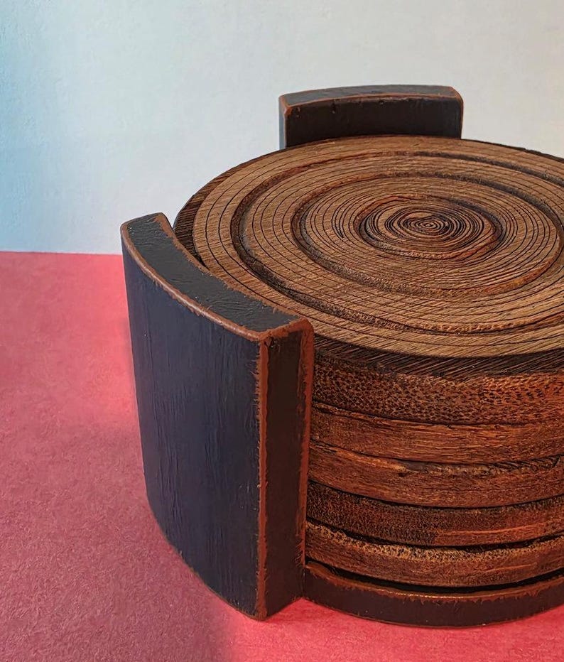 Pu&ograve; includere: Una pila di sottobicchieri circolari in legno con venature visibili, inseriti in un supporto in legno nero con finitura opaca. I sottobicchieri presentano anelli concentrici che ricordano i cerchi di crescita degli alberi.