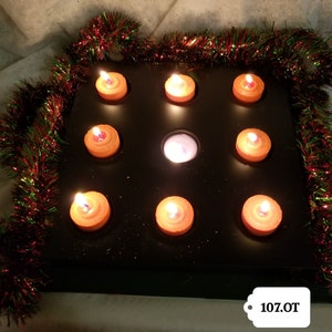 May include: A black metal tray with nine small orange candles, some lit, arranged in a grid pattern. The tray is decorated with green and red tinsel.