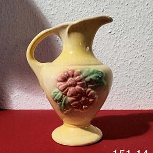 May include: A yellow ceramic pitcher with a handle and a spout. The pitcher features a raised floral design in pink and green. The base is a pedestal shape. The number "151.14" is printed at the bottom.