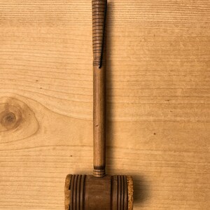 May include: A wooden gavel with a dark brown head and a lighter brown handle. The handle has carved grooves and a black cord for hanging. The gavel is displayed against a light brown wooden background.