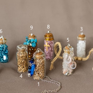 May include: A collection of small glass bottles filled with various decorative elements, including beads, shells, and crystals. Some bottles have cork stoppers and are adorned with chains or twine. The bottles are arranged on a neutral background.