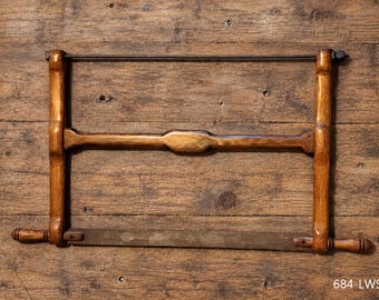 Può includere: Una sega a telaio in legno con una struttura tinta marrone scuro, una barra di supporto orizzontale centrale e una sottile lama in metallo fissata sul fondo. L'attrezzo include due piccole impugnature in legno sui lati e il testo 684-LWS1.