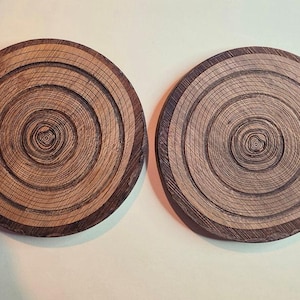 Pu&ograve; includere: Due sottobicchieri circolari in legno con un motivo a grana naturale in sezione trasversale. Ogni sottobicchiere mostra anelli concentrici in varie tonalit&agrave; di marrone chiaro e scuro, evidenziando la consistenza organica del legno.