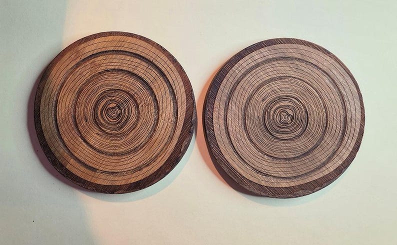Pu&ograve; includere: Due sottobicchieri circolari in legno con un motivo a grana naturale in sezione trasversale. Ogni sottobicchiere mostra anelli concentrici in varie tonalit&agrave; di marrone chiaro e scuro, evidenziando la consistenza organica del legno.