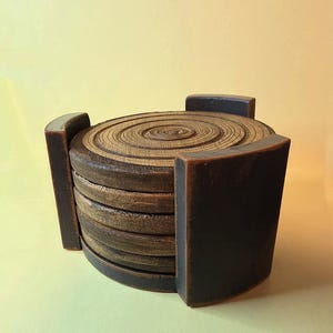 Pu&ograve; includere: Una pila di sottobicchieri circolari in legno con un motivo a venature di anelli di crescita sulla superficie superiore. I sottobicchieri sono tenuti in posizione da un supporto in legno curvo marrone scuro con tre supporti verticali.