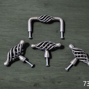 May include: A collection of silver-toned cabinet hardware, including a handle and three knobs, arranged on a dark green wooden surface. The hardware features a decorative, woven wire design. The number "738-3-" is visible in the bottom right corner.