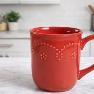May include: A red ceramic mug with a curved handle. The mug has a decorative dotted pattern near the rim and a smooth, glossy finish. The mug is on a white marble surface. The text "713-CS" is on the right side of the mug.