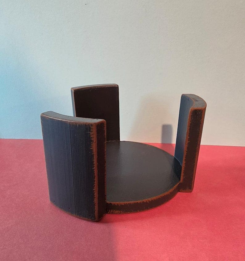 Pu&ograve; includere: Un portasottobicchieri in legno marrone scuro con base circolare e tre supporti verticali curvi. I bordi del legno presentano una finitura usurata, che rivela tonalit&agrave; marrone chiaro lungo gli angoli e i bordi della struttura.