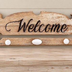 May include: A rectangular wooden wall sign with a distressed white paint finish over natural wood. The word Welcome is written in black cursive script. The sign features a horizontal cutout slot and three white knobs for hanging items, with 216-BWS2.
