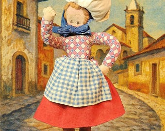 Può includere: Bambola di stoffa vintage vestita con abiti tradizionali, che porta un sacco bianco sulla testa. La bambola indossa un top a quadri blu e rossi, una gonna a quadri blu e bianchi e una gonna rossa. Lo sfondo raffigura una scena di villaggio europeo. Il numero "683-12" è in basso.