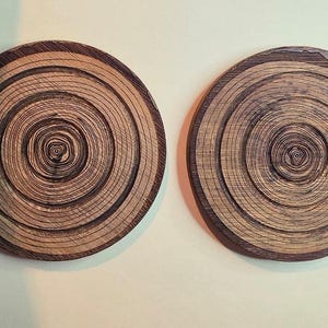 Pu&ograve; includere: Due sottobicchieri circolari in legno con un motivo naturale ad anelli di crescita. La superficie di ogni sottobicchiere mostra anelli concentrici di venature del legno marrone scuro e marrone chiaro, creando una texture dettagliata che imita il tronco.