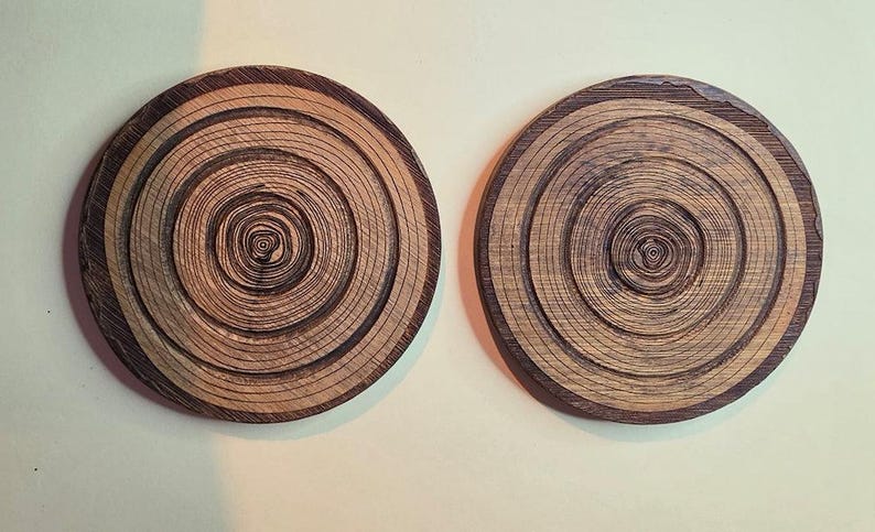 Pu&ograve; includere: Due sottobicchieri circolari in legno con un motivo naturale ad anelli di crescita. La superficie di ogni sottobicchiere mostra anelli concentrici di venature del legno marrone scuro e marrone chiaro, creando una texture dettagliata che imita il tronco.
