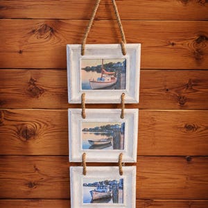 May include: A vertical hanging wall display featuring three rectangular white wooden frames connected by thick jute rope. Each frame contains a photograph of a boat docked in calm water. The top frame shows a sailboat, the middle a small rowboat, and the bottom a fishing vessel. The number 60- is printed in the bottom right corner.