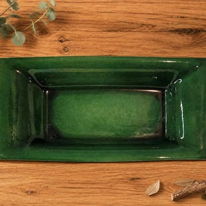 May include: A rectangular ceramic tray with a glossy, deep emerald green glaze. The dish features a smooth, reflective surface with subtle variations in color intensity, including darker tones near the base and edges, and a wide, flat rim.