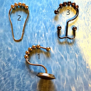 May include: Three shower curtain hooks with decorative ball accents. The hooks are in different styles and finishes, including gold and bronze tones. The hooks are displayed on a blue surface with a water-like pattern.
