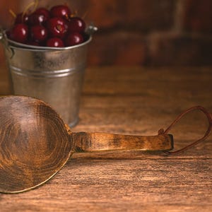 May include: A rustic wooden scoop with a long handle and a leather loop, next to a small metal bucket filled with ripe red cherries. The scoop has a dark, textured finish, and the scene is set on a wooden surface.