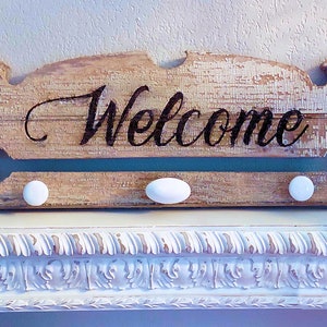 May include: A rustic wooden sign with the word "Welcome" painted in black lettering. The sign is mounted on a white shelf with decorative trim and the number "216" painted on the front.