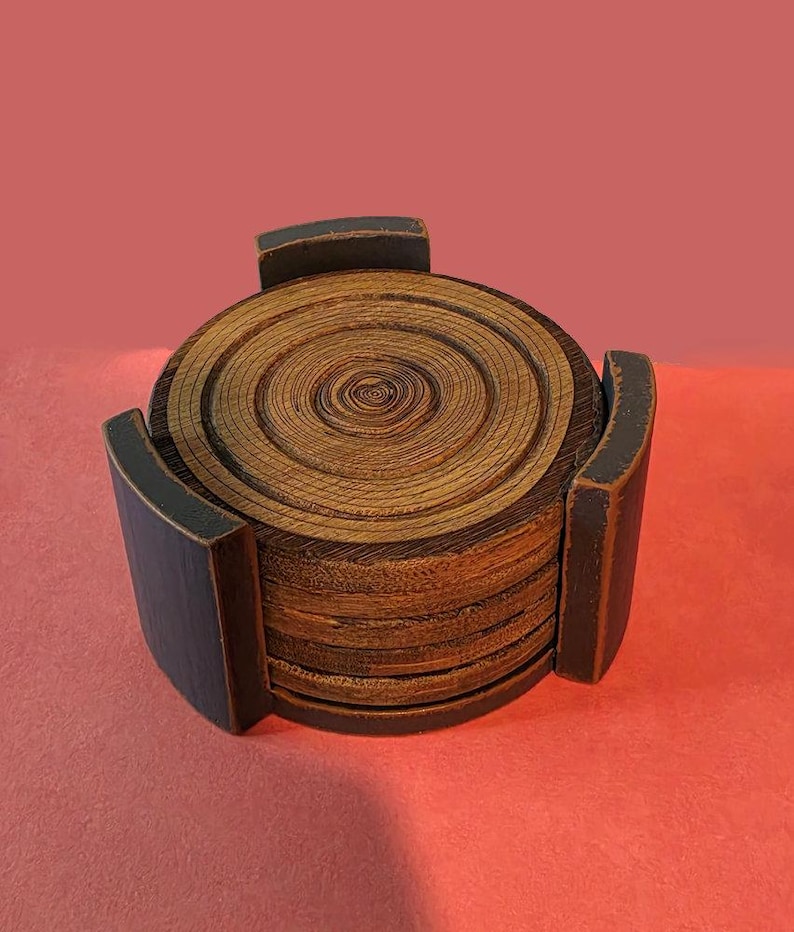 Pu&ograve; includere: Una pila di sottobicchieri circolari in legno con venature visibili, inseriti in un supporto in legno nero con finitura opaca. I sottobicchieri presentano anelli concentrici che ricordano gli anelli di crescita degli alberi.