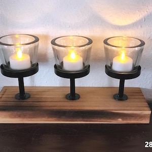May include: A wooden candle holder with three glass votives, each holding a white LED candle. The votives are supported by black metal stands on a rectangular wooden base. The overall design is rustic and decorative.