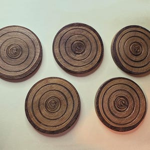 Pu&ograve; includere: Un set di cinque sottobicchieri circolari in legno, ognuno caratterizzato da una venatura naturale che imita gli anelli concentrici di un tronco d'albero. I sottobicchieri sono marrone scuro con accenti beige chiaro e hanno un design piatto.