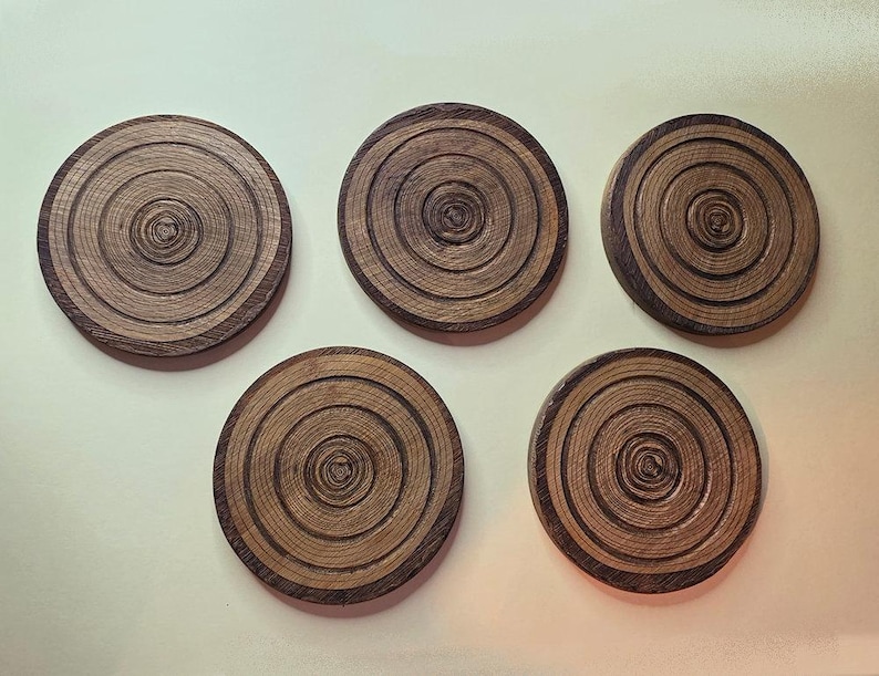 Pu&ograve; includere: Un set di cinque sottobicchieri circolari in legno, ognuno caratterizzato da una venatura naturale che imita gli anelli concentrici di un tronco d'albero. I sottobicchieri sono marrone scuro con accenti beige chiaro e hanno un design piatto.