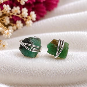 May include: A pair of stud earrings featuring raw, uncut green emerald stones wrapped in silver-colored wire. Each stone displays natural variations in color and texture, with the wire coiled securely around the center of the rough gemstone.