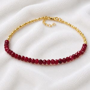 May include: A delicate bracelet featuring a strand of small, faceted, deep red gemstone beads that transition into a chain of tiny, polished gold-tone metal beads. The piece includes a gold-tone lobster claw clasp and an adjustable extender chain.