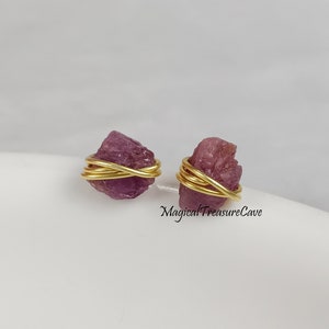 May include: A pair of raw, pink gemstone stud earrings. Each stone is wrapped with gold-colored wire. The earrings are displayed on a white surface. The text "Magical Treasure Cave" is visible.