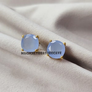 以下が含まれることがあります： ゴールドカラーの爪が付いた、ライトブルーの宝石スタッドピアス。 ピアスは丸く、ファセット加工されており、画像には「MAGICALTREASURECAVE」の文字が見えます。