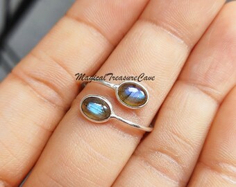 Può includere: Un anello d'argento con due pietre ovali di labradorite. L'anello è aperto e regolabile per adattarsi a diverse dimensioni delle dita. Le pietre preziose hanno un bagliore iridescente blu e verde.