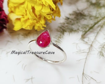 Puede incluir: Un anillo de plata con una piedra preciosa de color rosa intenso, tallada en forma de pera y montada en una banda metálica sencilla. El texto MagicalTreasureCave está impreso en negro sobre la superficie junto al anillo. Diseño limpio.