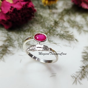 May include: A silver ring featuring a vibrant oval-cut ruby gemstone. The ring has a feather design and the text "Magical Treasure Cave". The ring is set against a white surface with floral accents.