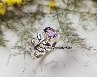 May include: A silver ring featuring an oval, light purple gemstone. The ring has a leaf-like design on one side and the text "Magical Treasure Cave" is visible. The ring is set against a white background with greenery.