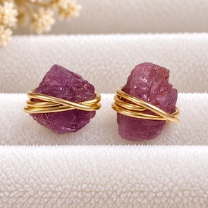 May include: A pair of stud earrings featuring raw, deep pink gemstone nuggets secured by polished gold-tone wire wraps. The stones have an irregular, textured surface and are held in place by multiple loops of thin, metallic gold wire.