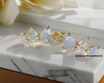 May include: Four gold rings with iridescent, pear-shaped moonstone gemstones. The rings are displayed on a white marble surface. The text "MAGICALTREASURECAVE" is visible in the lower right corner.
