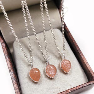 May include: Three leaf-shaped pendants with silver chains. The pendants are a light orange color and have etched leaf vein details. The necklaces are displayed inside a brown jewelry box with a light gray interior.