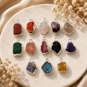May include: A collection of twelve raw gemstone pendants, each featuring a rough, irregular stone encased in a silver-toned metal border with a small circular jump ring at the top. The stones display a variety of colors including red, orange, white, dark purple, clear, green, pink, black, violet, blue, and yellow.