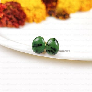 Peut inclure: Une paire de boucles d'oreilles clous en pierre précieuse verte et noire avec des tiges dorées. Les boucles d'oreilles sont sur une surface blanche.