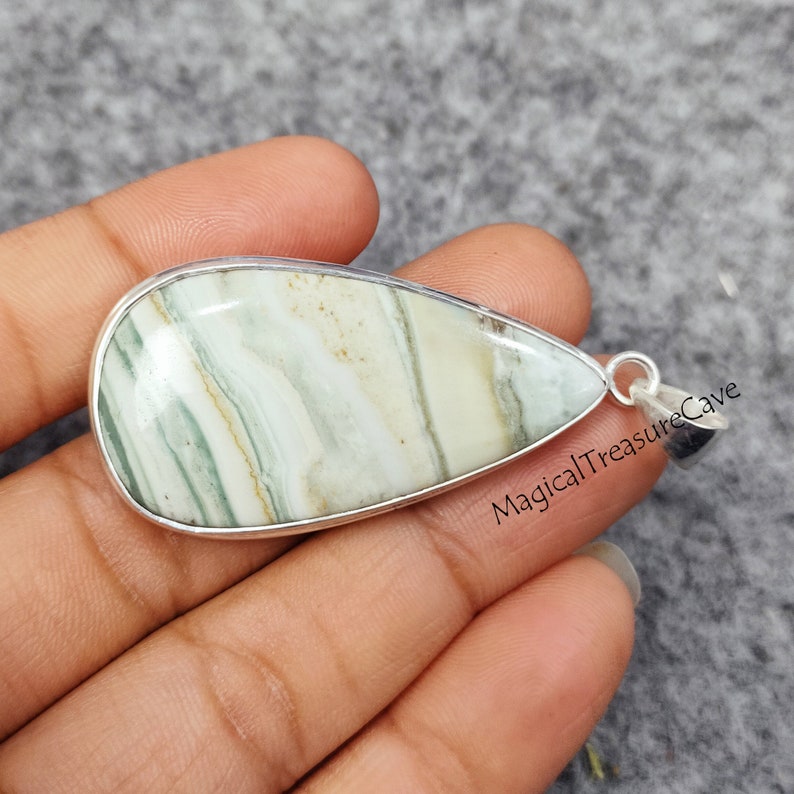 May include: A teardrop-shaped pendant with a green and white agate stone set in a silver frame. The pendant is on a hand, and the text "MagicalTreasureCave" is visible in the background.