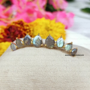 May include: A pair of gold-toned ear climbers featuring teardrop-shaped labradorite stones. The earrings have a curved design with multiple stones, showcasing a mix of gray, blue, and green hues. The jewelry is displayed on a neutral surface.