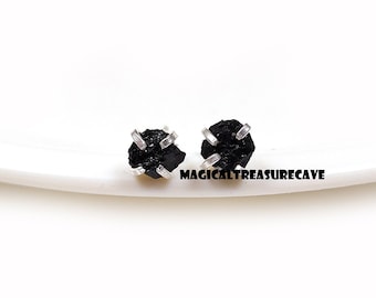 May include: A pair of black tourmaline stud earrings. Each earring features a rough, natural-looking black stone held in place by a silver-colored metal setting. The earrings are displayed on a white surface. The text "MAGICALTREASURECAVE" is visible.