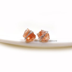 Op de afbeelding: Een paar oorbellen met kleine, ruwe, oranje sunstone-kristallen die zijn gewikkeld in zilverdraad. De oorbellen liggen op een wit oppervlak.