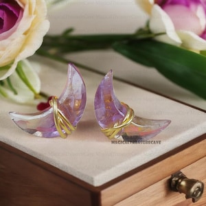 Può includere: Un paio di orecchini a forma di mezzaluna. Gli orecchini sono realizzati in materiale trasparente e iridescente con una tonalità viola. Sono avvolti con filo color oro. Sono esposti su una scatola di legno.