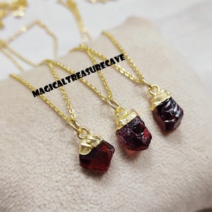 Puede incluir: Tres collares dorados con colgantes de granate rojo oscuro en bruto. Los colgantes tienen formas irregulares y tapas doradas. Los collares se exhiben sobre una superficie beige claro. El texto "MAGICALTREASURECAVE" es visible.
