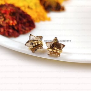 Puede incluir: Un par de pendientes en forma de estrella. Cada pendiente presenta una estrella de cristal de cuarzo ahumado facetado, envuelta con alambre dorado. Los pendientes se exhiben sobre una superficie blanca, con el texto "Magicaltreasurecave" visible.