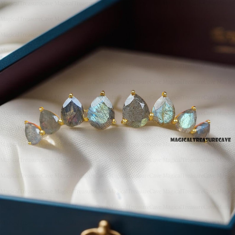 May include: A pair of gold-toned earrings featuring teardrop-shaped labradorite stones. The stones display iridescent blue and green hues. The earrings are presented in a blue jewelry box with a white satin lining. The text "MAGICALTREASURECAVE" is visible.